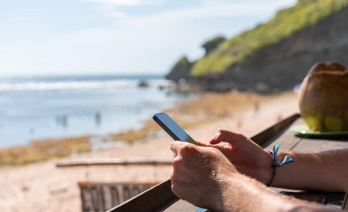 Close up on a man's hands holding a cell phone with a coconut and beach in the background