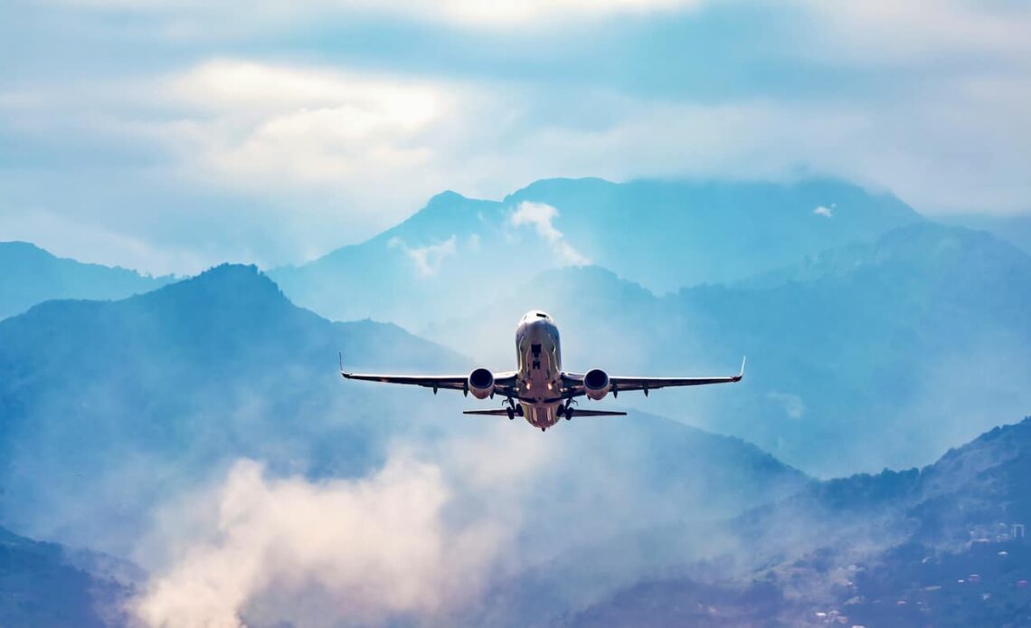 A lone commercial jet flying through a bright blue sky with mountains in the distance