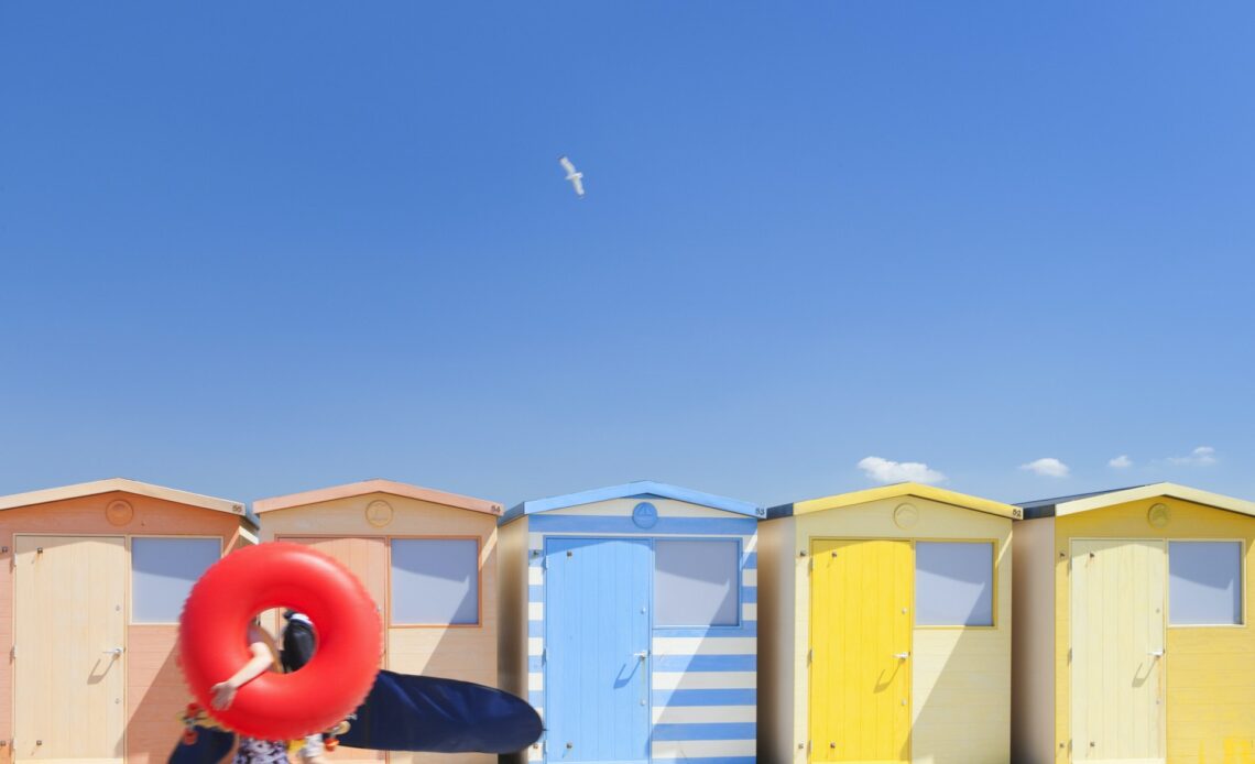 People carrying inflatables walk past colorful beach huts on the English coast