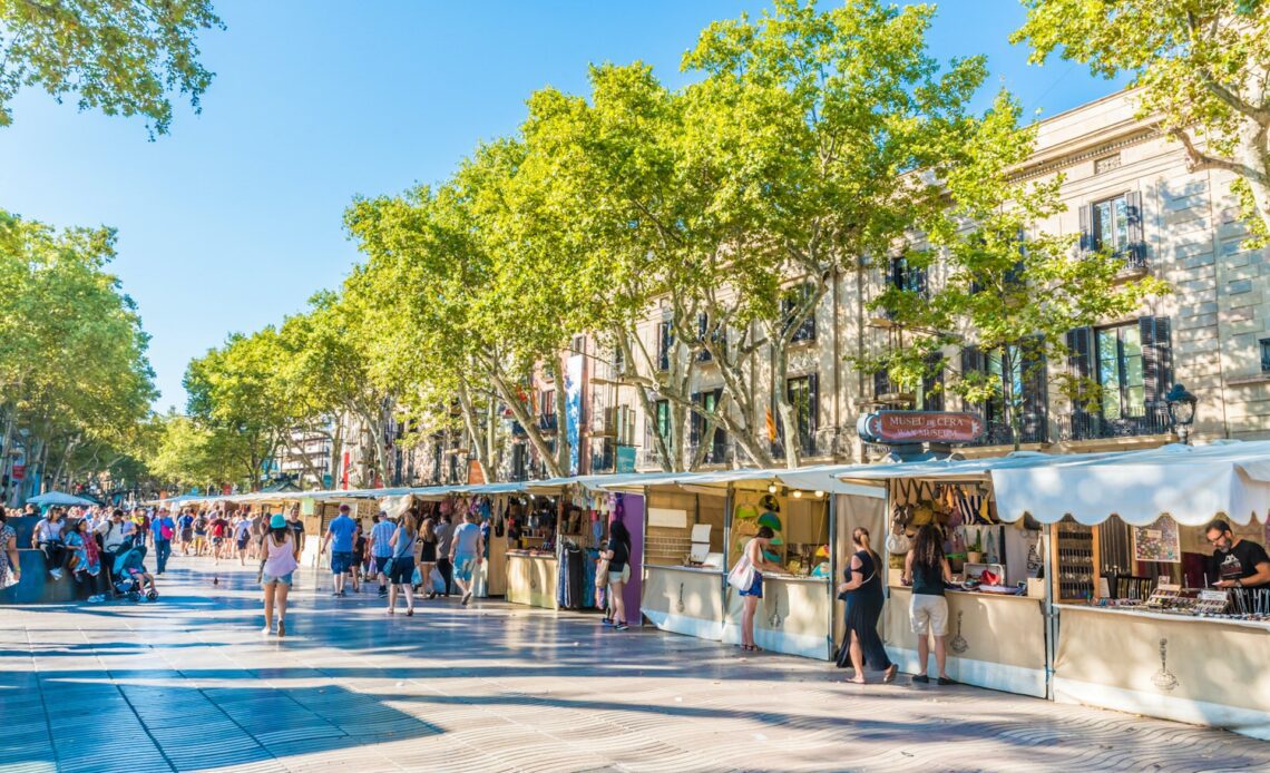 Bright blue skies over shoppers browsing market stalls on tree-lined La Rambla: one of the essential free things to do in Barcelona