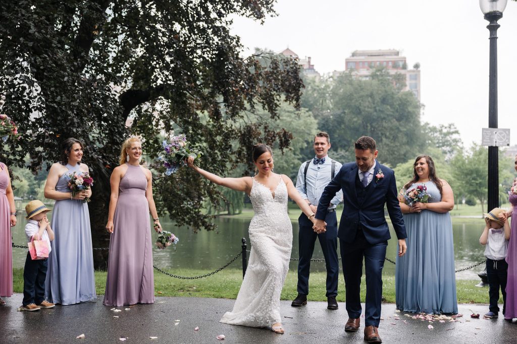 Kate and Charlie after their Boston wedding ceremony, Kate holds up her bouquet with one arm, holding Charlie's hand, walking down the "aisle" as bridesmaids look on and cheer.