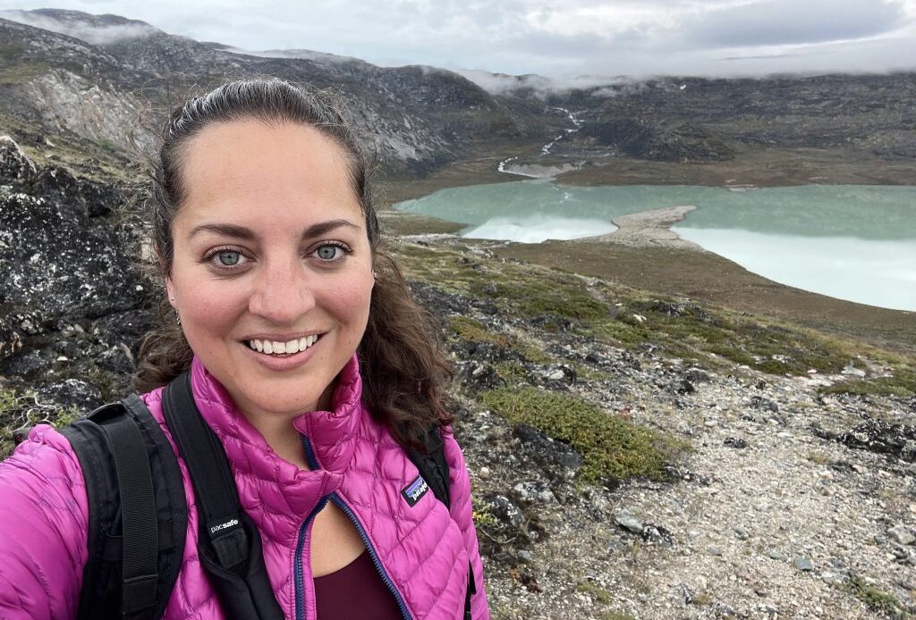 Kate taking a selfie wearing a pink jacket and hiking near a still green lake in Greenland.