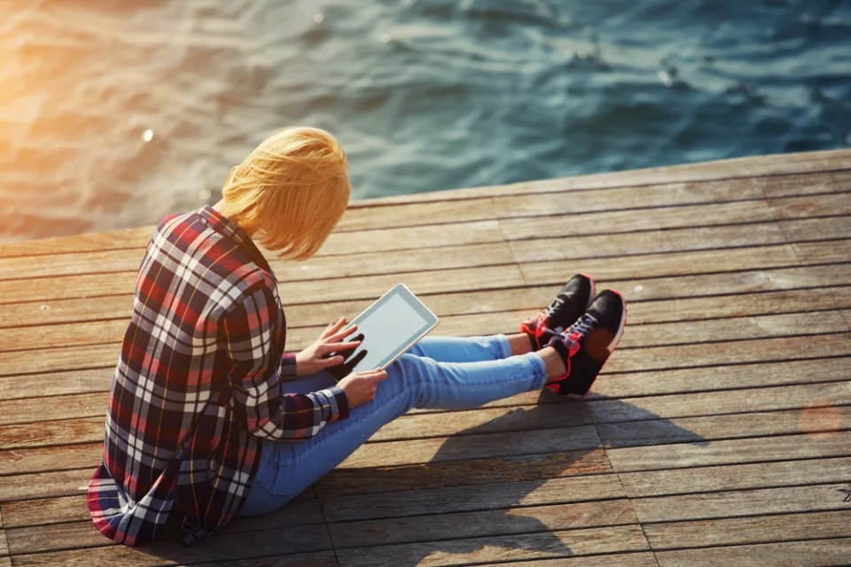 Side view of blonde hair woman using digital tablet while sitting on a wooden pier next to the sea, female tourist busy use tablet while sitting outdoors at sunny day, girl browsing outside, flare sun