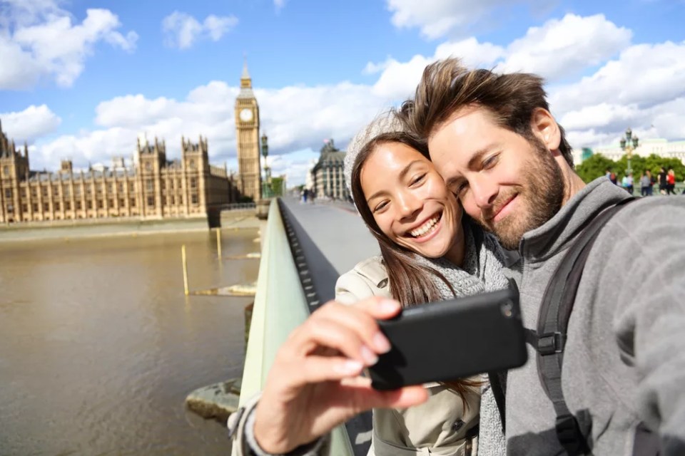 London tourist couple taking photo near Big Ben. Sightseeing woman and man having fun using smartphone camera smiling happy near Palace of Westminster, Westminster Bridge, London, England.