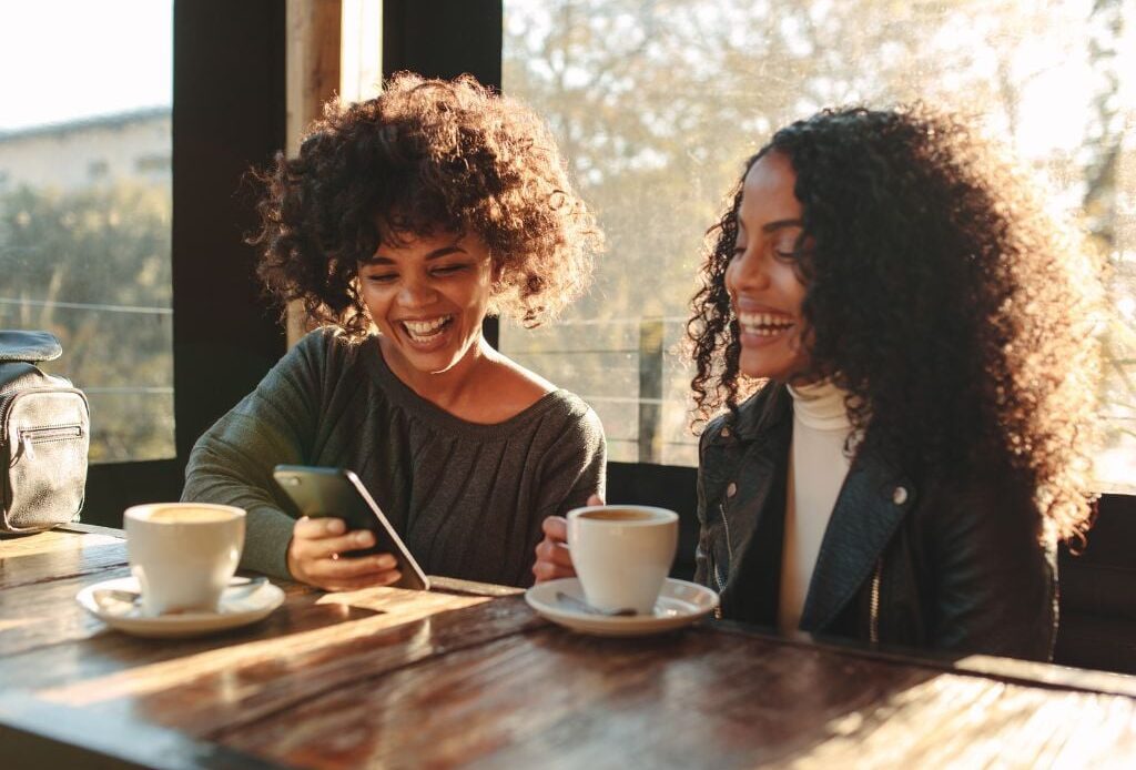 two women sitting at a table looking at an iphone with coffee cups