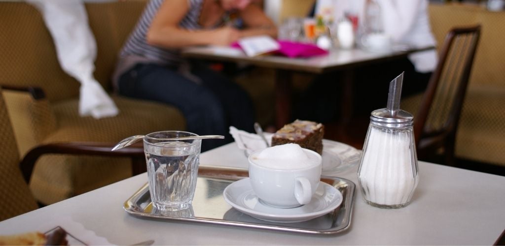 A cup of coffee, glass of water, a battle of sugar and a slice of cake in a small plate on the white table.
