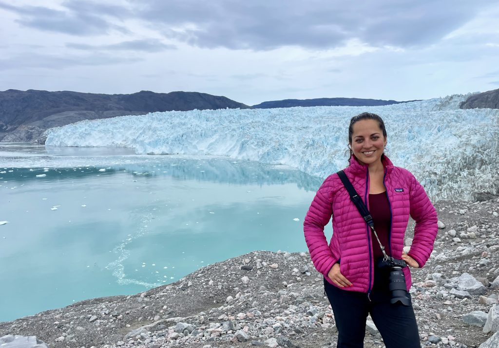 Kate wearing a bright pink jacket and a big camera, standing in front of a massive blue-white glacier on a still bay.
