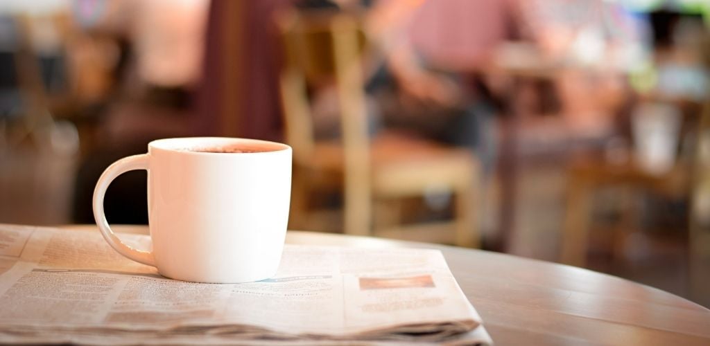On the table there is a white cup of hot coffee on the news paper 