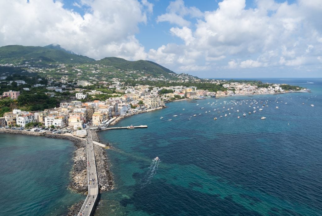 A view of the jagged coastline of Ischia, lots of little white boats in the bright turquoise water, and a bridge leading from the island to the castle.