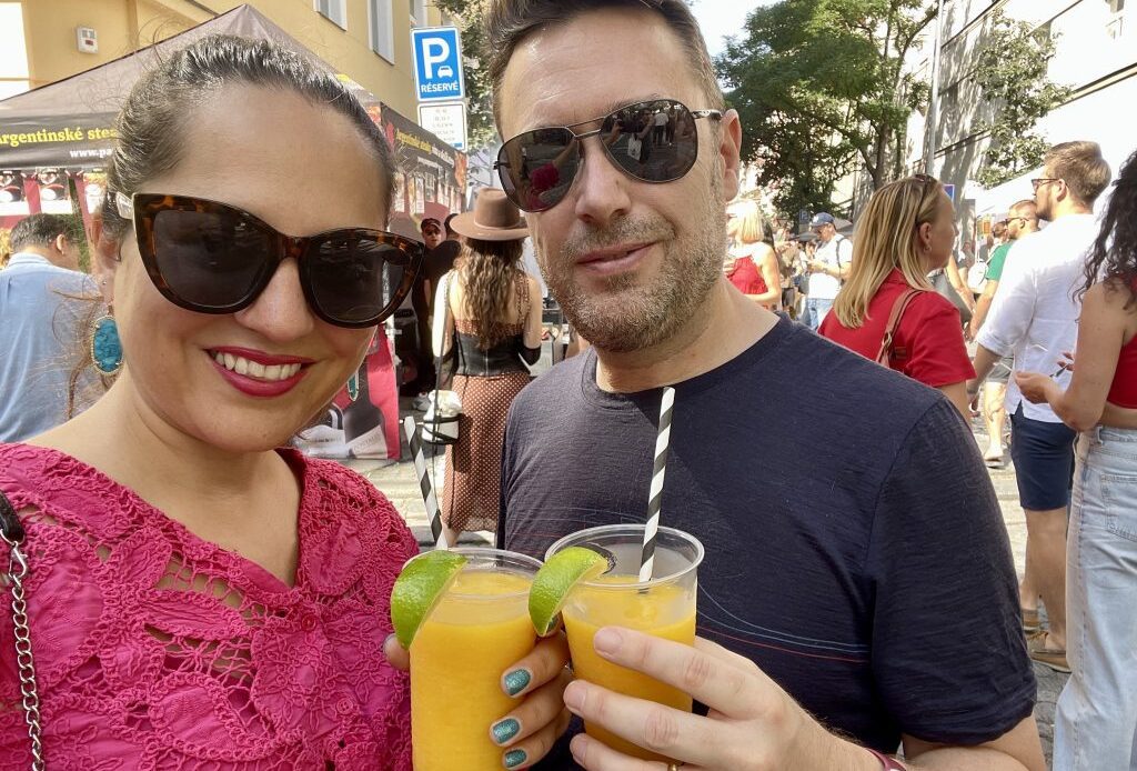 Kate and Charlie taking a smiling selfie in sunglasses, each holding a frozen mango margarita, with a street festival in the background.