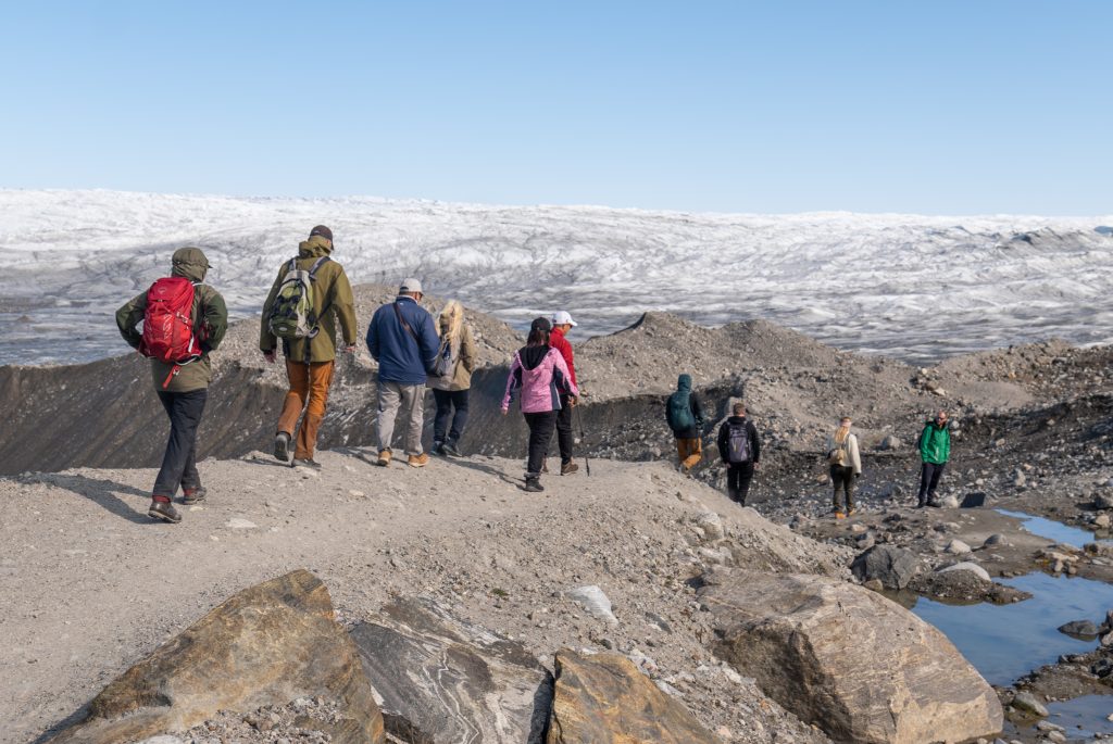 A line of people hiking along the ridge of a rock, a big glacier in the distance.