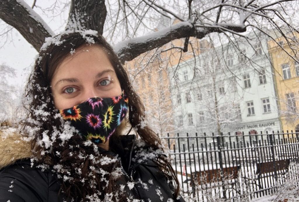 Kate poses for a selfie in a snowstorm with giant flakes in her hair and on her black jacket. She wears a black face mask with multicolor hearts on it and smiles. Behind her are a tree covered with snow, some park benches, and pastel orange, green, and yellow homes in the distance.