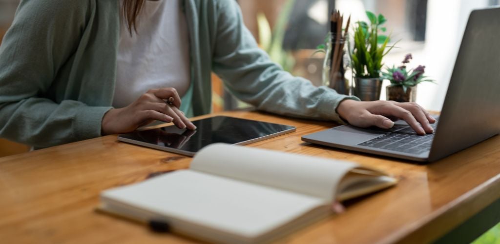 A woman writing making list taking notes in notepad and digital tablet working or learning on laptop indoors-educational course and there is notebook on the side of the table.