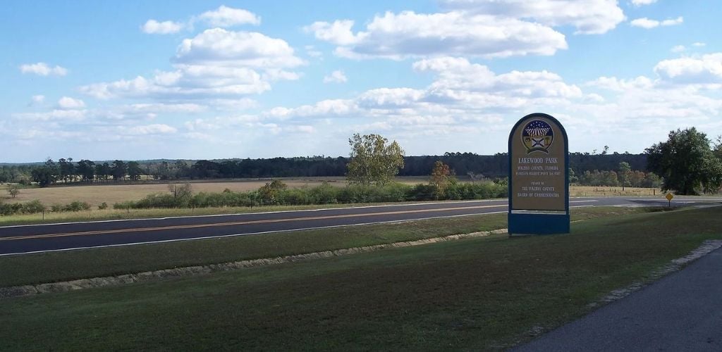 A long road and a signage of Lakewood park surrounded by green field land and trees. 