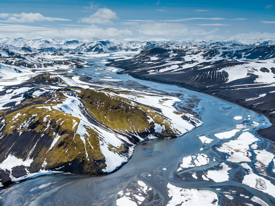 Glacial river stream winding through black volcanic sand, green moss and white snow