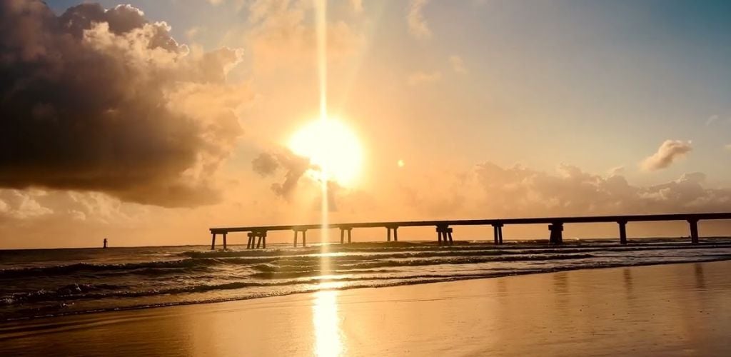 A beach with a long bridge that leads into the sea at sunset.