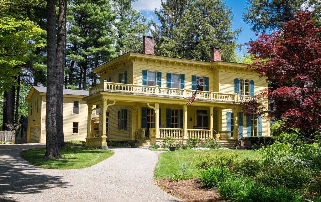An old-fashioned yellow house with dark green shutters and a large porch set among trees at the end of a driveway in Stockbridge, Massachusetts.