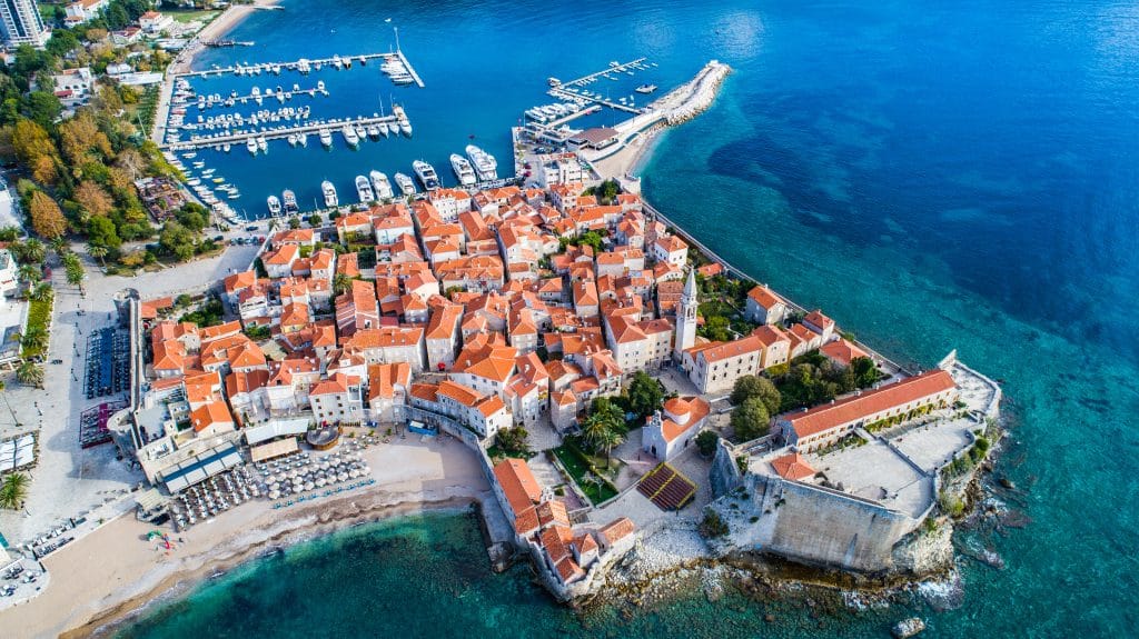 A tiny walled city with orange roofs surrounded by the bright blue ocean and beaches on each side.