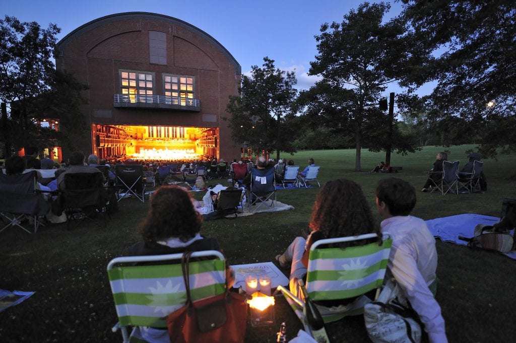 People sitting on folding chairs on grass outside Tanglewood Music Hall in Lenox, Mass.