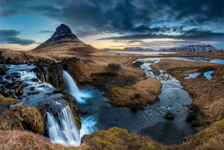 waterfall spilling over cliff into pool with cone shaped mountain behind it in Iceland