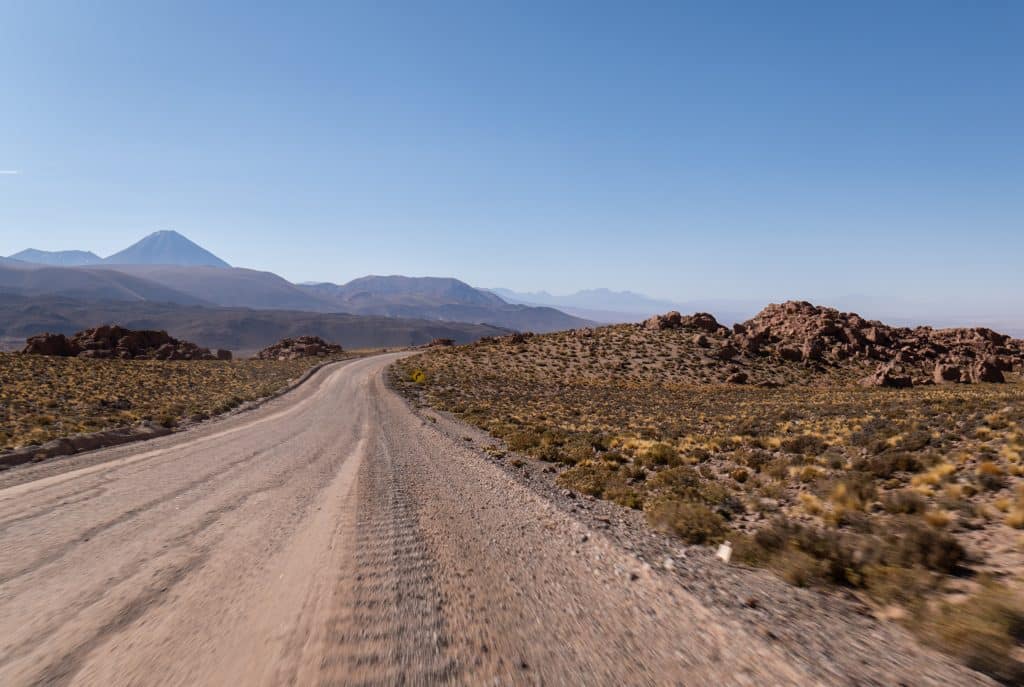 A dirt road leading through the Atacama Desert, with purple-blue mountains in the distance, surrounded by bright orange rocky hills covered with green shrubs.