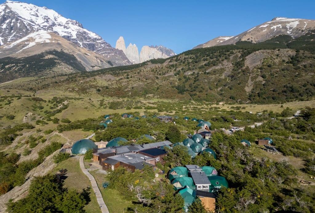 A drone view of Ecocamp Patagonia -- a cluster of small green domes in the middle of a hilly, grassy area, with a snowy mountain and the three jagged gray rock towers in the distance behind it.