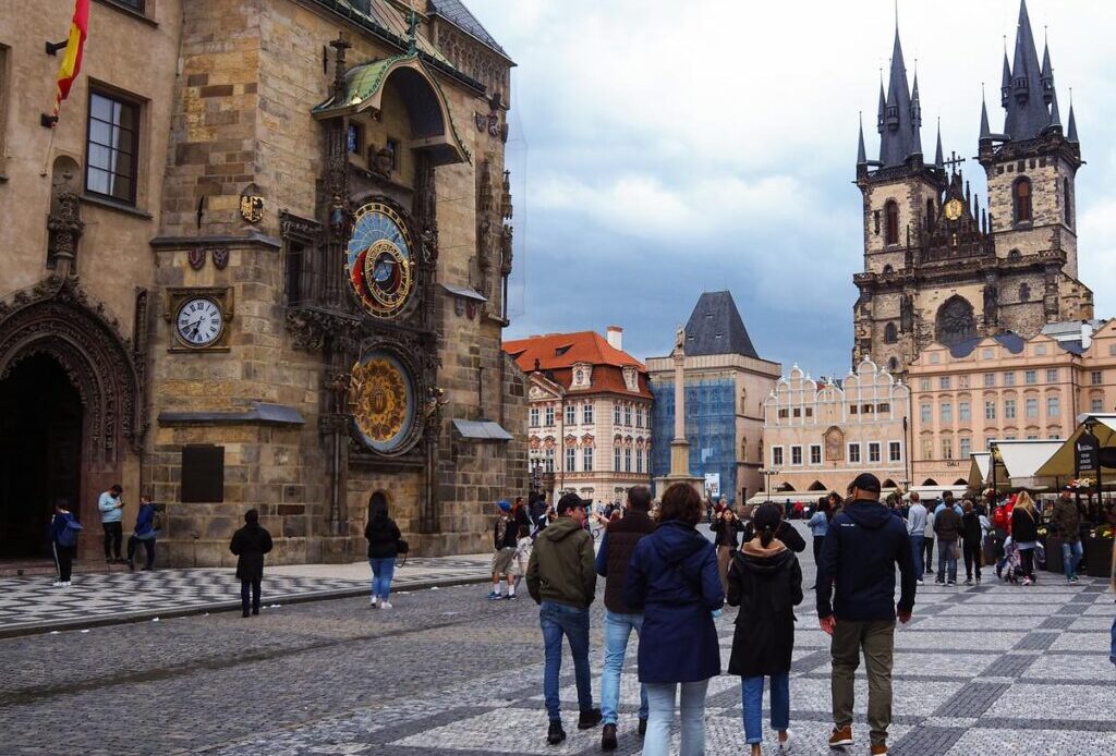 People walking and gathering in Prague’s Old Town Square with the Prague Astronomical Clock and the Church of Our Lady before Týn in the background.