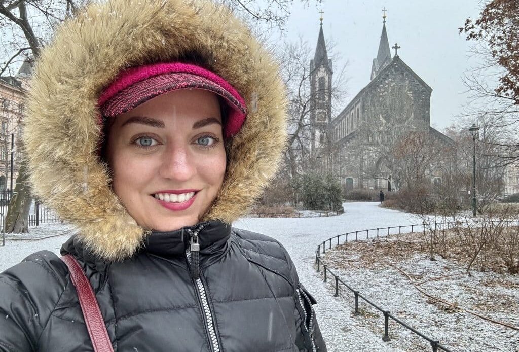Kate taking a smiling selfie wearing a big winter coat with a furry hood. She's on a snowy square in Prague, a Gothic church behind her.