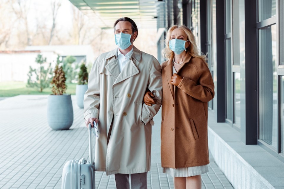 Mature business couple in medical masks standing near suitcase on urban street