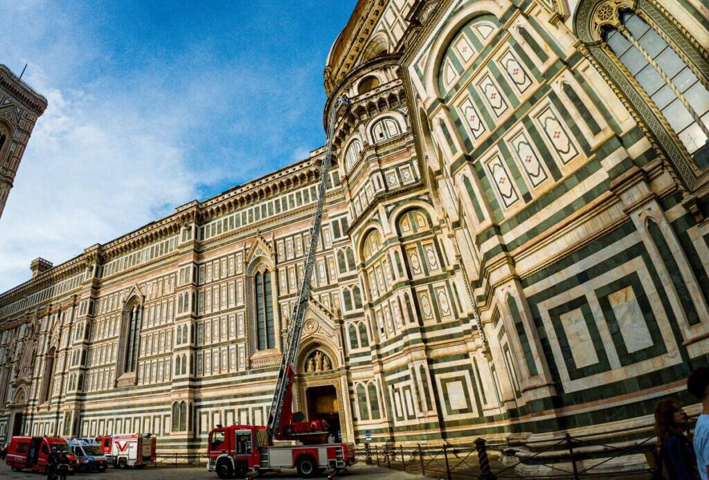 An emergency response team with a red fire truck and extended ladder at the Santa Maria del Fiore Cathedral, capturing a juxtaposition of modern intervention at a historic Italian landmark under a bright blue sky.