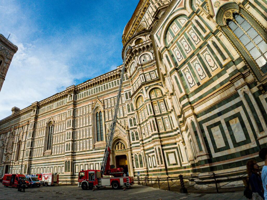 An emergency response team with a red fire truck and extended ladder at the Santa Maria del Fiore Cathedral, capturing a juxtaposition of modern intervention at a historic Italian landmark under a bright blue sky.