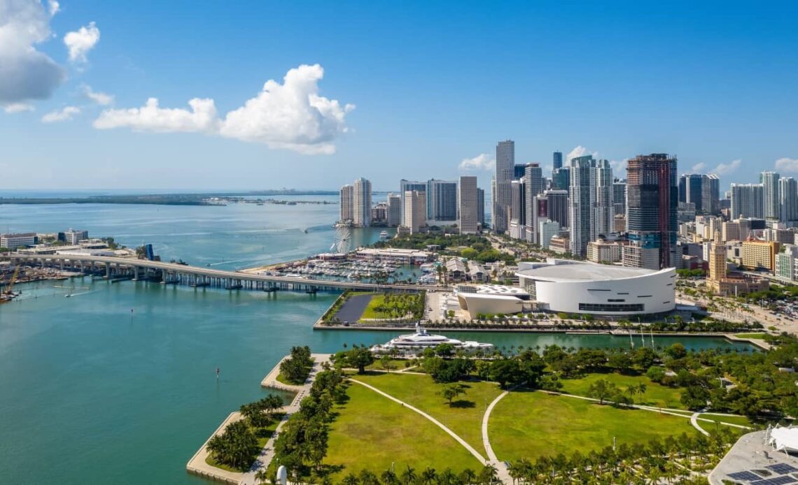 Aerial shot of Downtown Miami with its many skyscrapers, with a sprawling park in the foreground and a long bridge going to an island underneath a blue sky