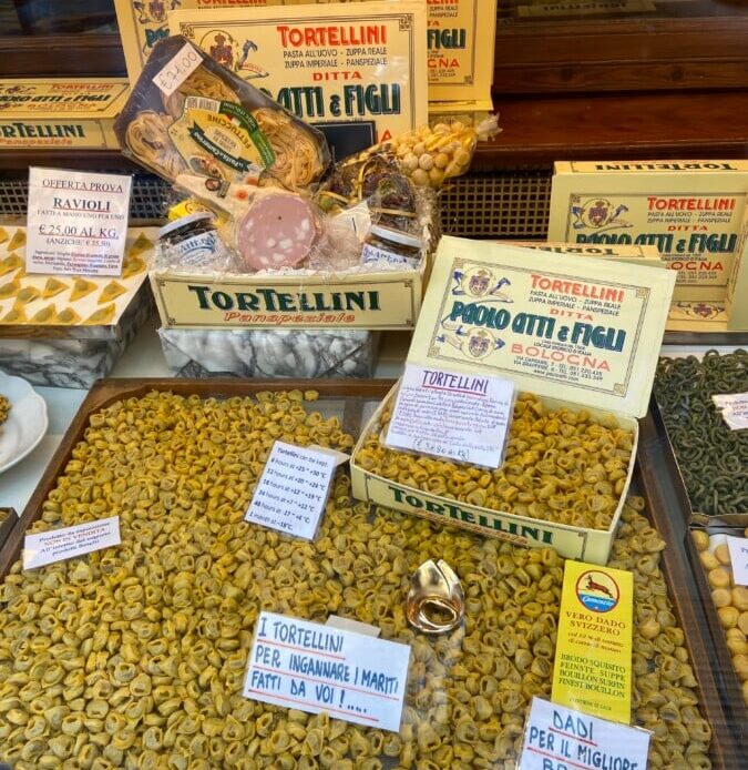 Window display of a traditional food shop in Bologna, Italy featuring mortadella, tortellini, and other pastas