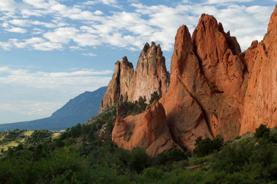 Blue sky and clouds over the Garden of the Gods rock formations in Colorado Springs, Colorado. The bushes and vegetation are a lush green against the red rock formations