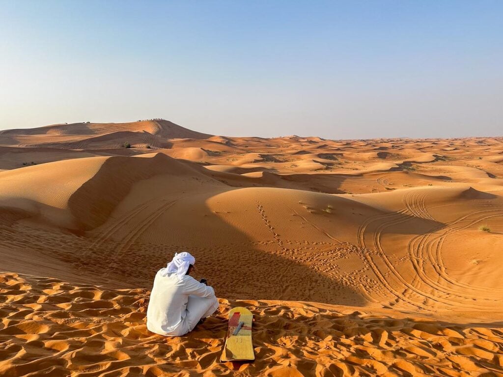 A sandboarder in traditional white clothing observes the endless rolling dunes of Dubai's Red Sand Desert at dusk.