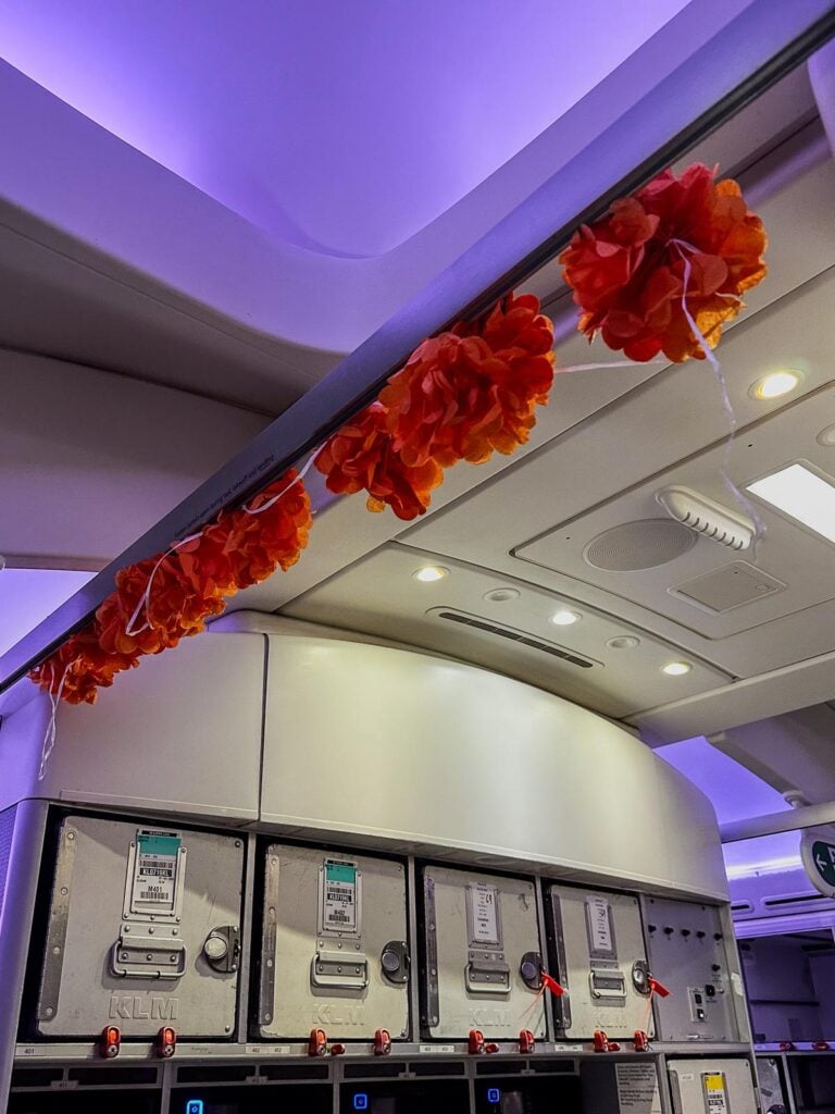 Orange garland decorations celebrating King's Day inside a KLM aircraft with violet lighting and branded overhead compartments.
