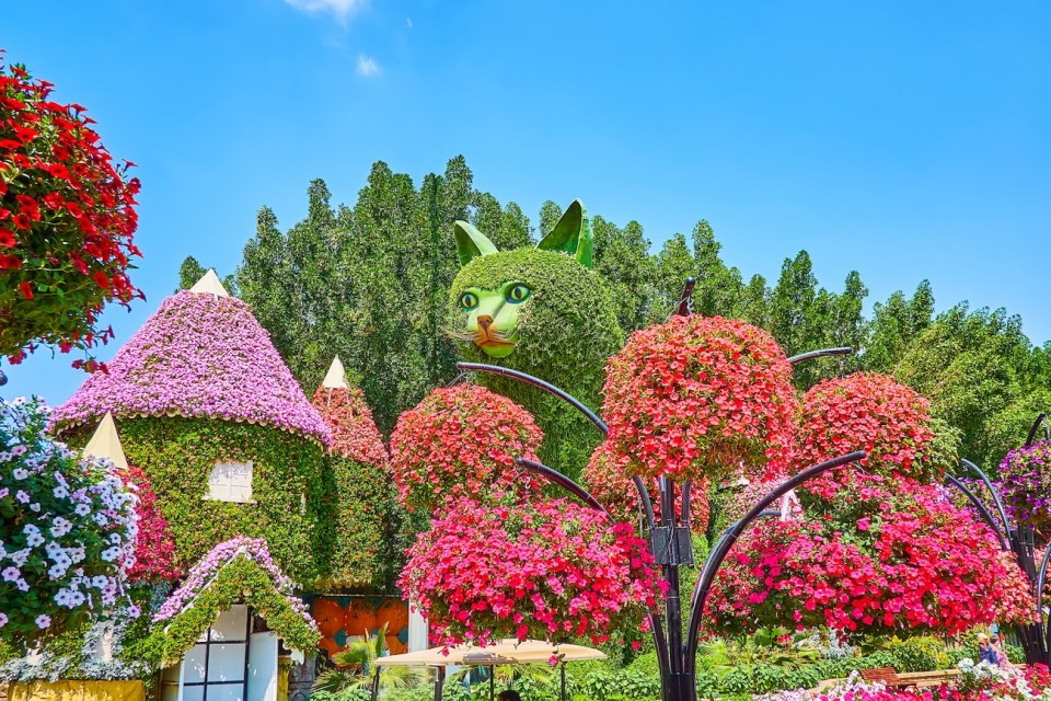 The hanging pots with bright petunia flowers in front of the installation of giant cat and house, covered with living plants, Miracle Garden