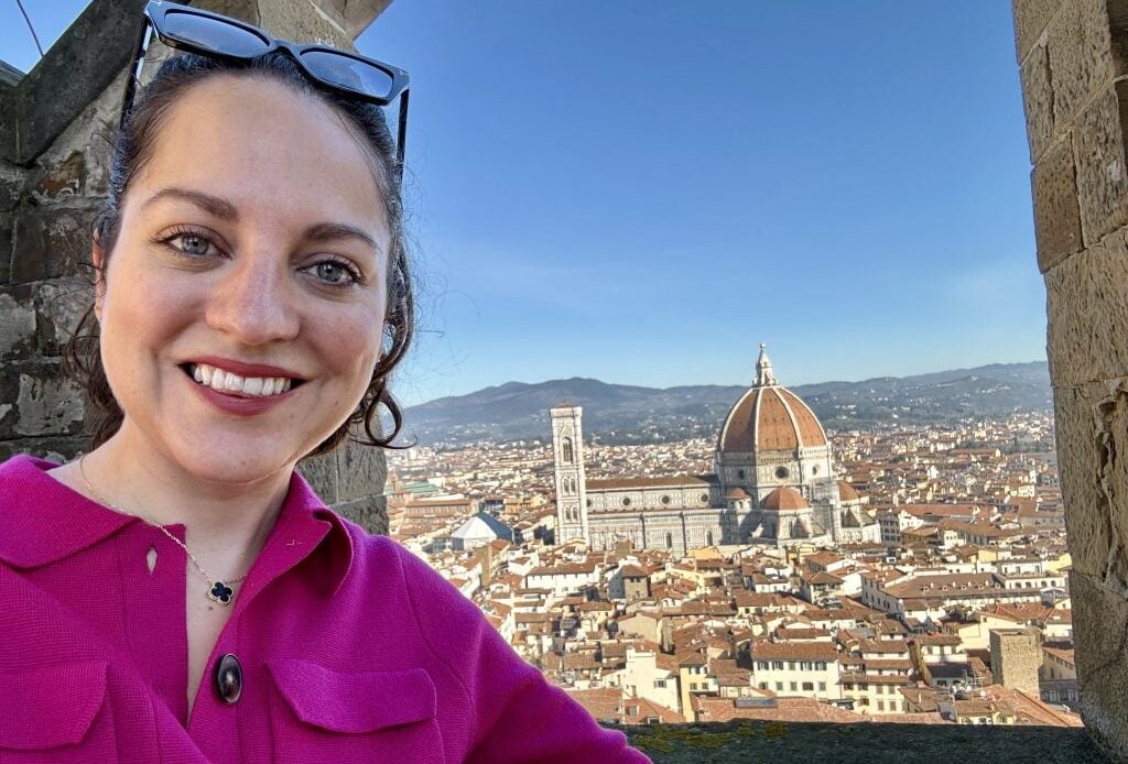 Kate taking a selfie in front of a window displaying the big Florence Duomo, surrounded by orange-roofed buildings.