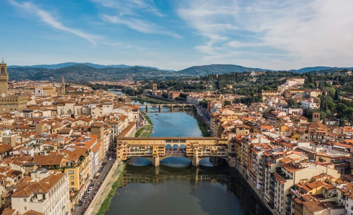 Aerial view of the city of Florence with the Arno River running through it, with Ponte Vecchio in the foreground and mountains in the background