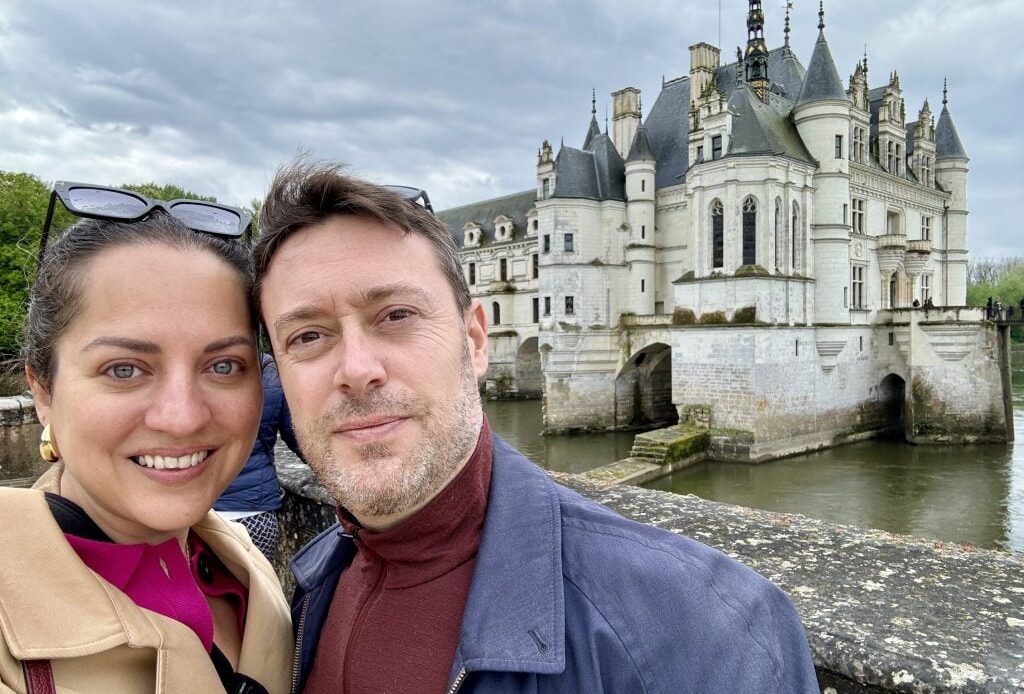 Kate and Charlie taking a smiling selfie in front of a big gray chateau perched on a river in France.