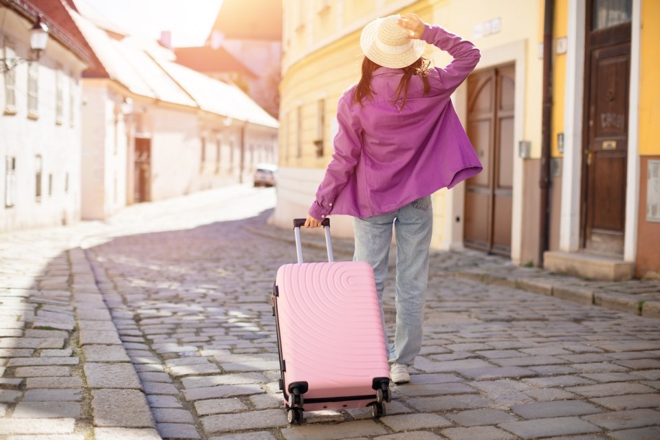 A woman in casual travel attire walks down a quaint cobblestone street in a picturesque European town, back view