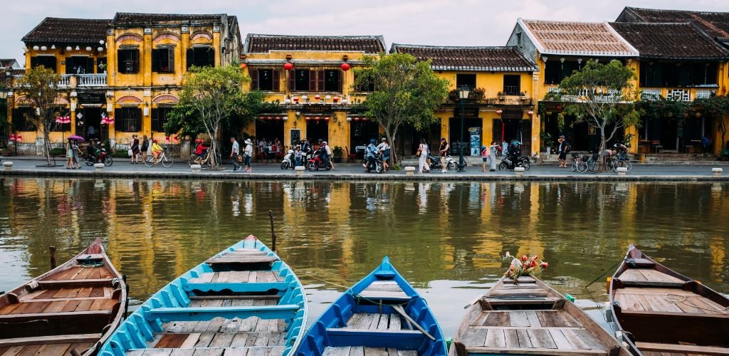 Hoi An, Vietnam, image of boats in foreground, river running through middle and traditional yellow painted buildings with people passing by