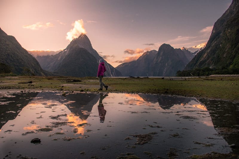person walking beside lake with mountains in background and refelcted in the water at sunset