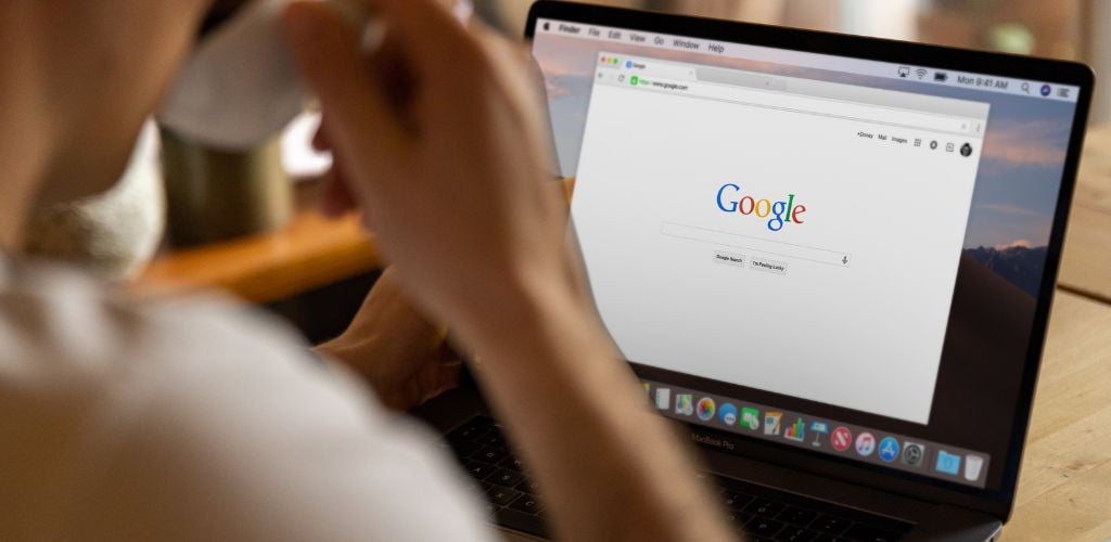 laptop open with Google search engine displayed on screen. Person drinking from a mug in front of desk in the foreground of image 