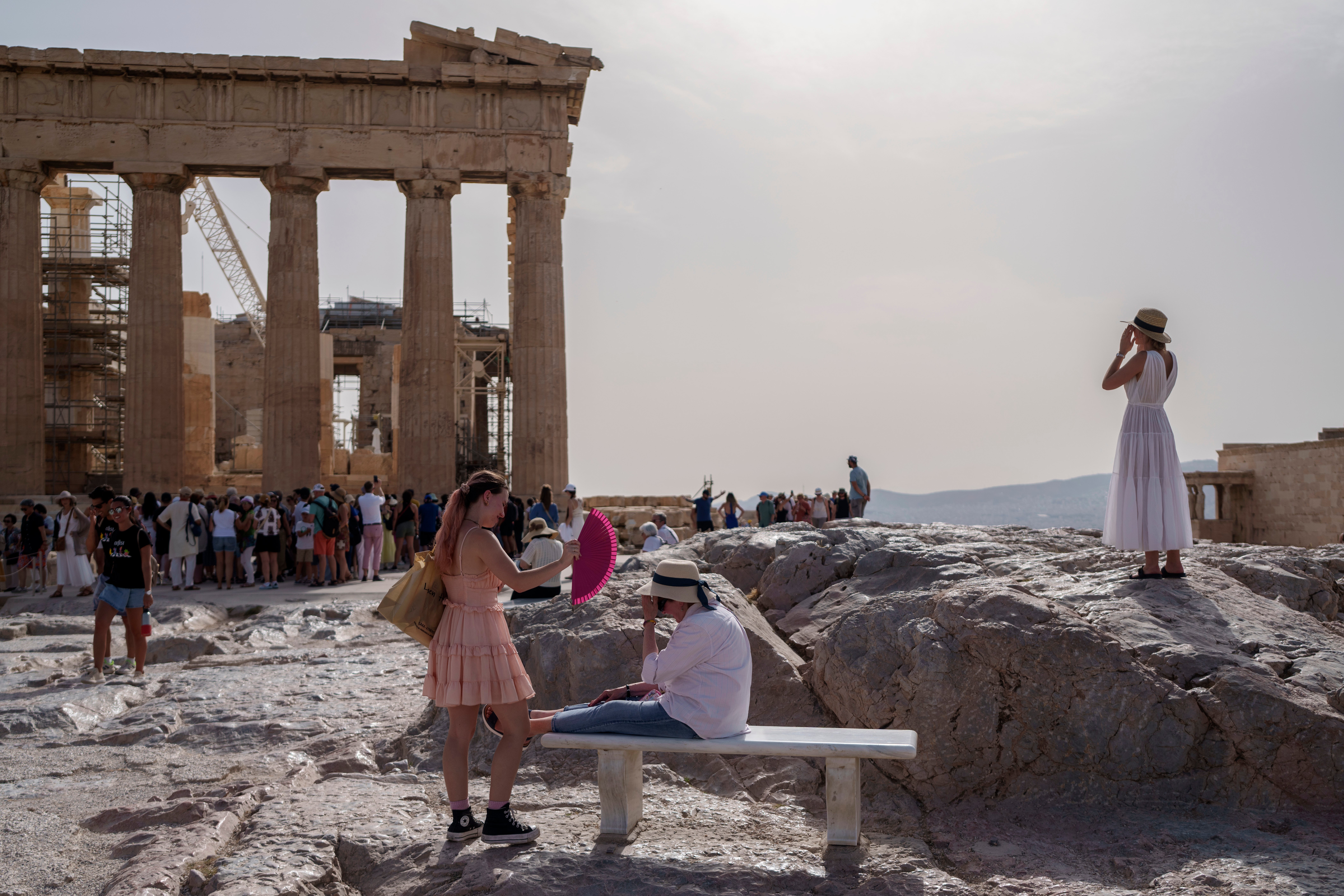 A tourist uses a hand fan to cool down another one sitting on a bench in front of the Parthenon at the ancient Acropolis, in Athens, Wednesday, June 12, 2024