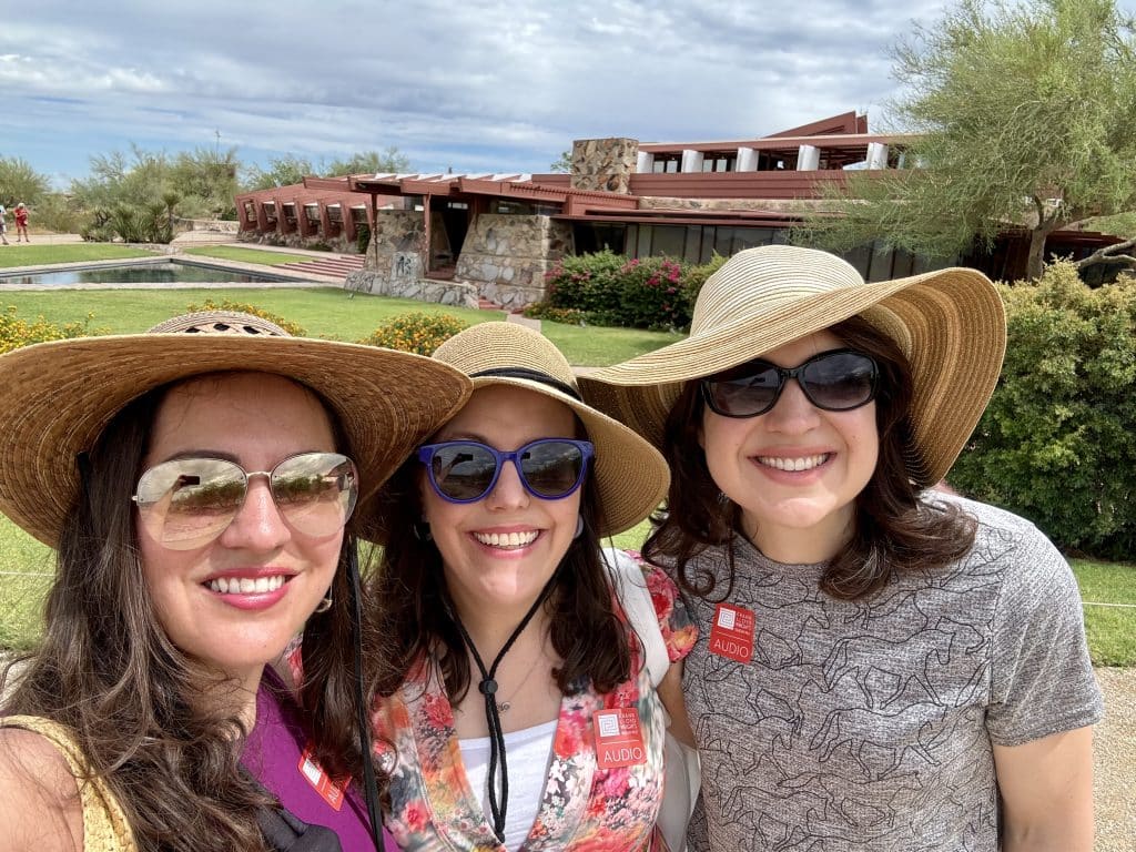 Kate and her two friends in floppy hats and sunglasses, with the modern red building of Taliesin West behind them.