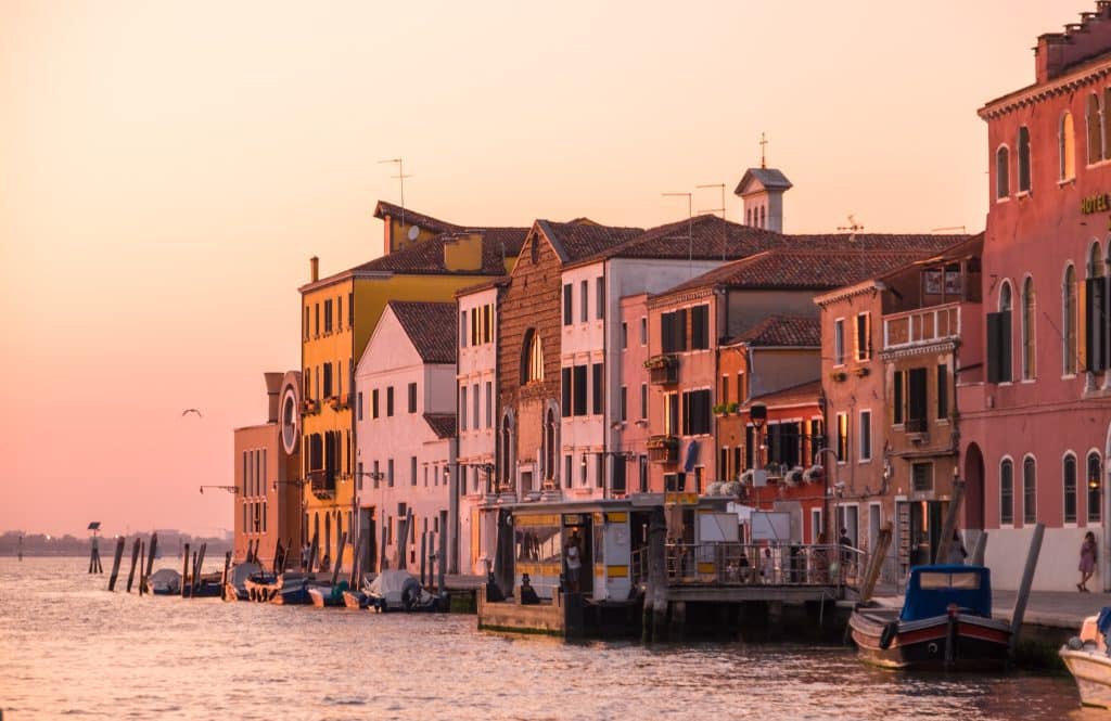 A row of houses in Venice against a canal, light up bright pink at sunset.