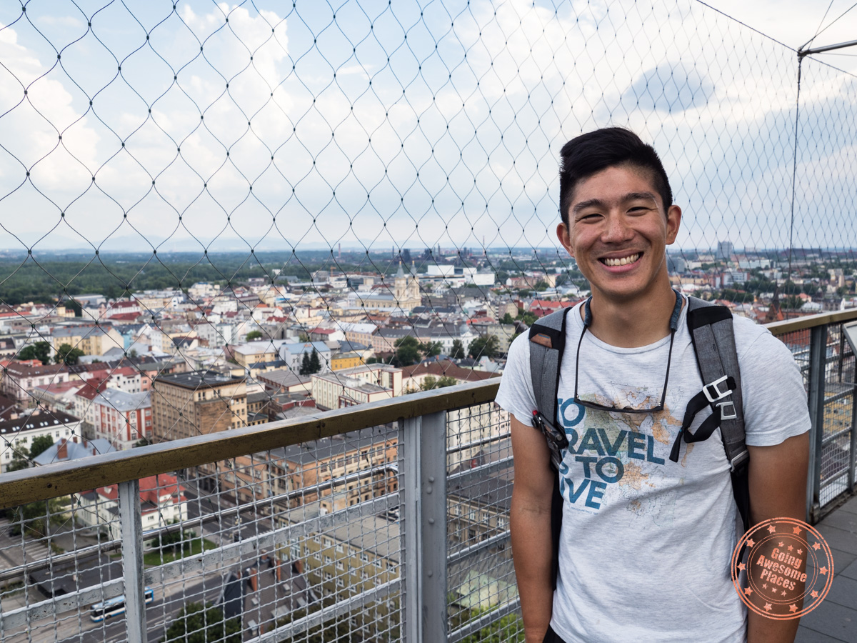 A smiling person stands on a rooftop observation deck with a cityscape view in the background. They are wearing a backpack and a T-shirt with text, and the area is enclosed with a safety fence. The scene appears to be during daylight, under a partly cloudy sky.