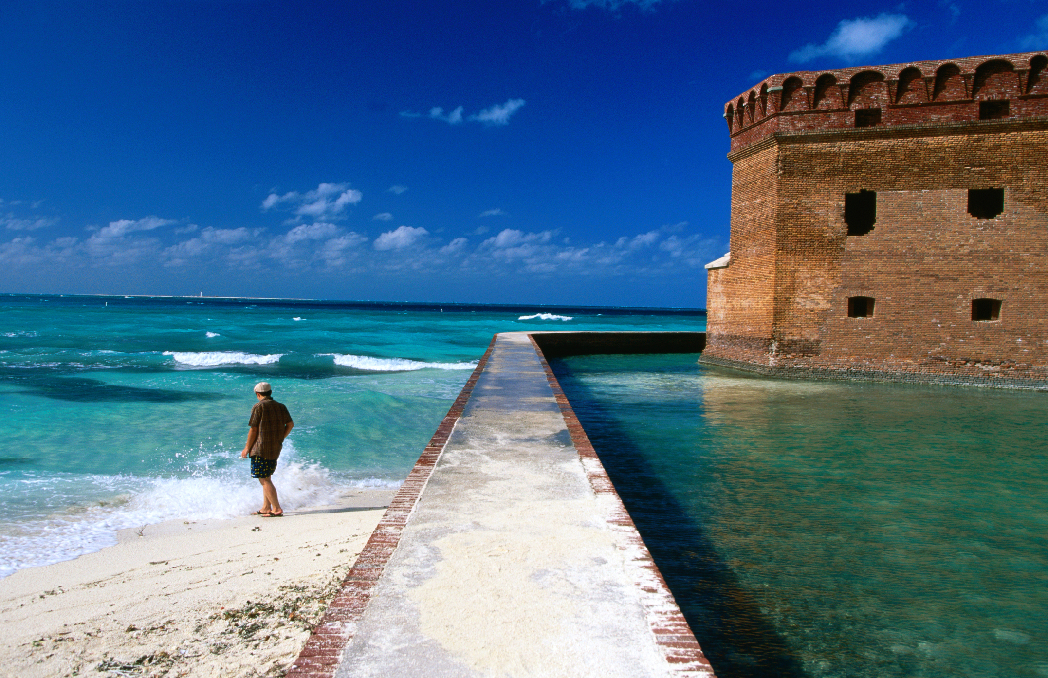 A solo figure dips his toes in the sea on a beach at the edge of a fortress