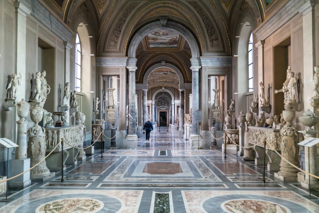 A museum in Rome with ornate floors, the walls covered with marble statues. A man with keys walks through the empty museum.